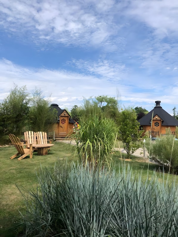 Une cabane en bois avec une table ronde et des chaises, située dans un espace herbeux, entourée de hautes herbes et d’arbres.