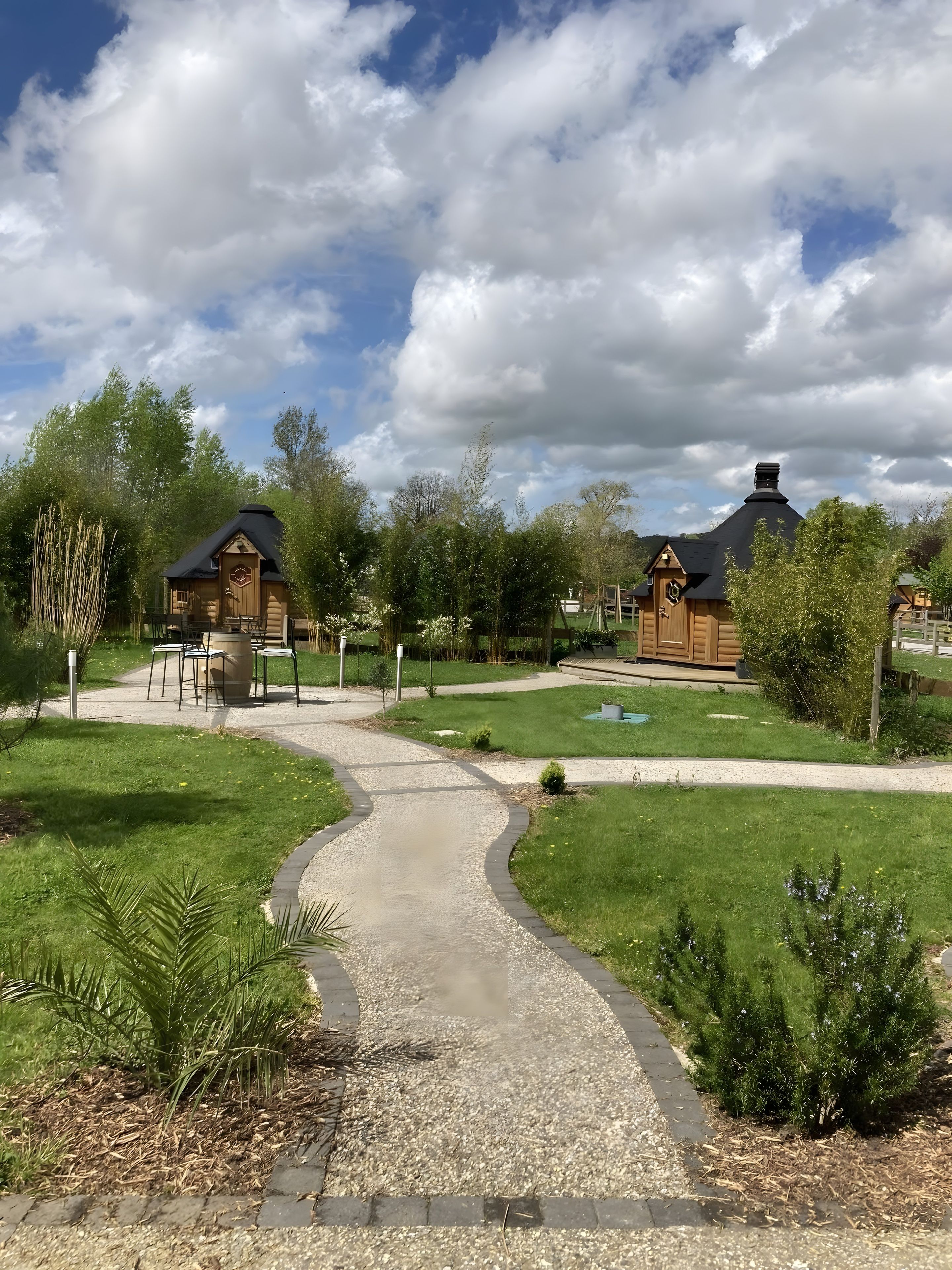 Un sentier mène à des cabanes en bois, avec une zone herbeuse et des arbres en arrière-plan.