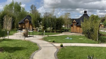 Un sentier mène à des cabanes en bois, avec une zone herbeuse et des arbres en arrière-plan.