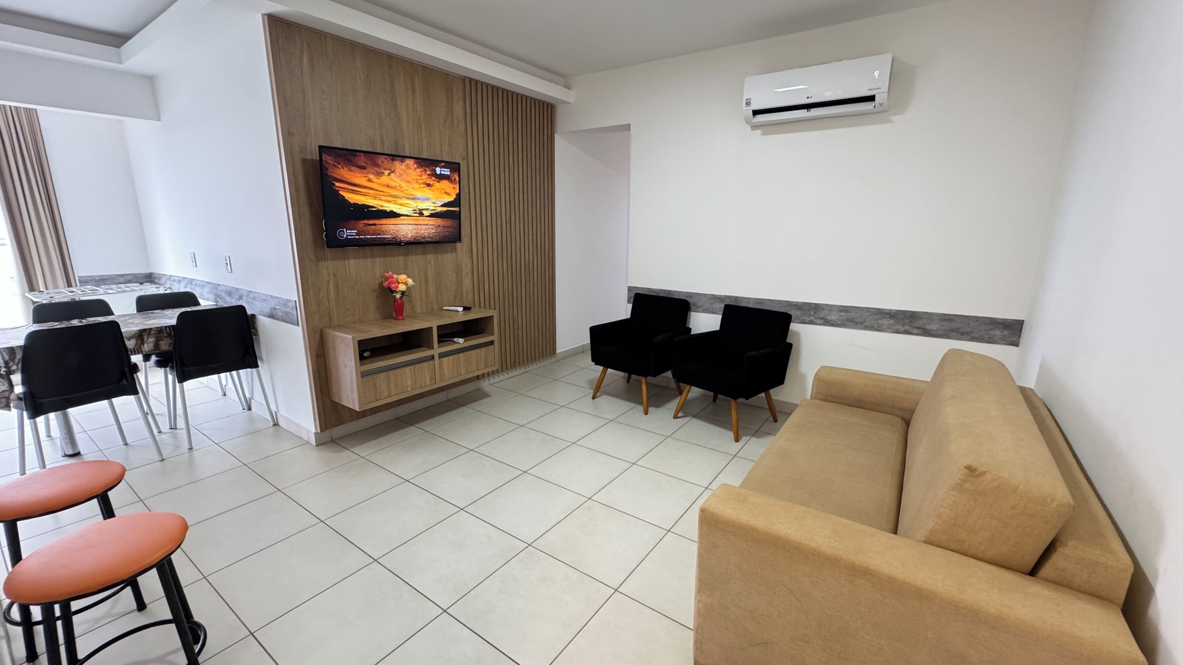 Uma sala de estar moderna com uma TV de tela plana, um sofá, duas cadeiras pretas, uma mesinha com um vaso, e uma área de jantar com cadeiras e uma mesa.