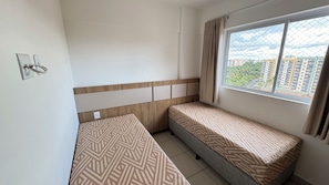 A small, clean room with two single beds, a window with a view of buildings, and a simple wall-mounted light switch. - CRCN - Evian Thermas Residence (Caldas Novas)