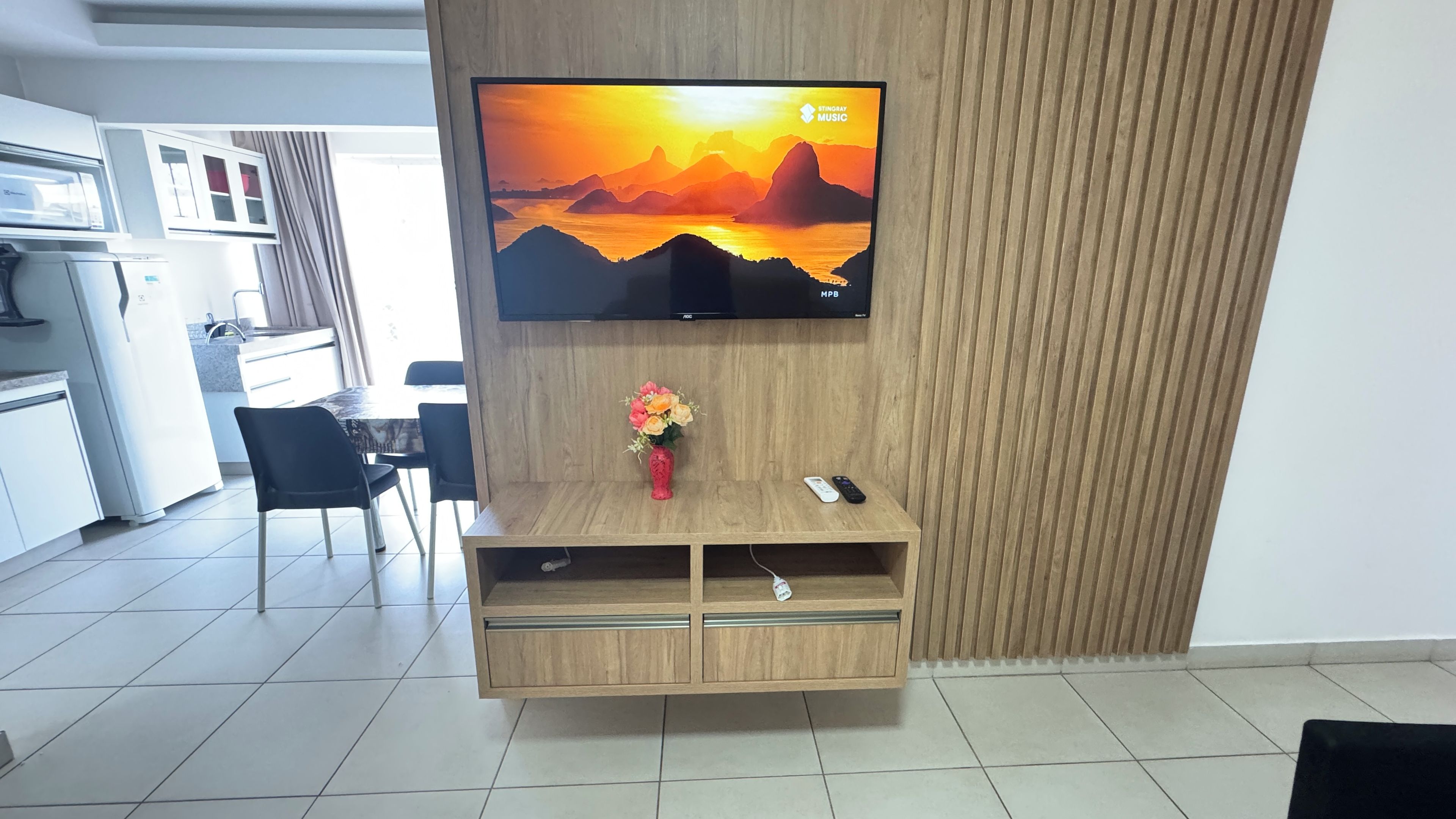 A modern living room with a flat-screen TV mounted on a wooden wall, a small wooden cabinet below, and a vase with flowers on the cabinet.