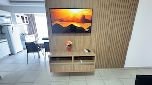 A modern living room with a flat-screen TV mounted on a wooden wall, a small wooden cabinet below, and a vase with flowers on the cabinet. - CRCN - Evian Thermas Residence (Caldas Novas)