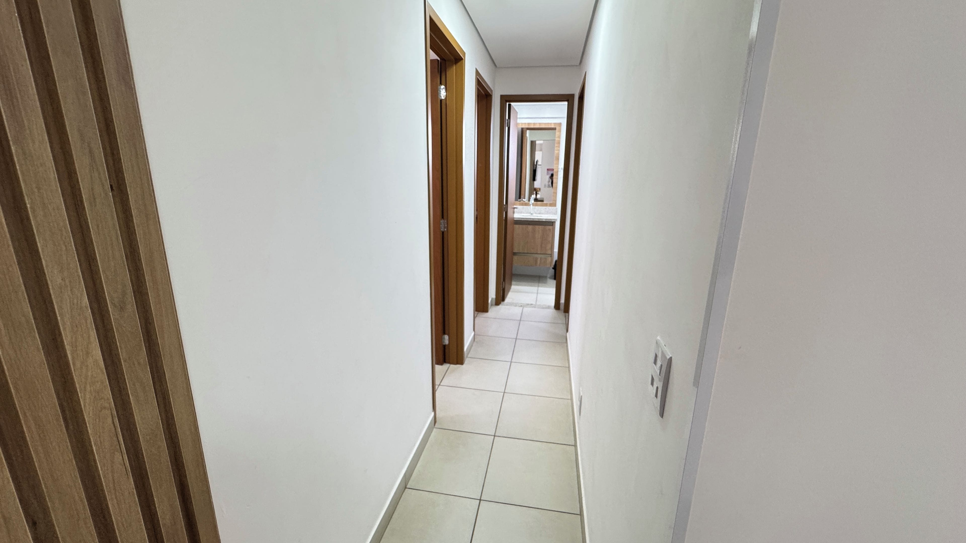 A narrow hallway with white walls and wooden doors, leading to a bathroom with a toilet and sink.