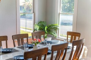 A dining table set for six with chairs, a vase with flowers, and a view of a street through large windows. - NEW! Fall in a Victorian | Grand Porches Near DT (San Antonio)