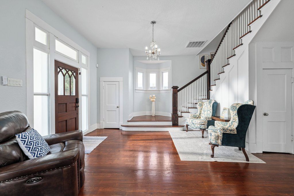 A spacious foyer with a wooden staircase, leather sofa, patterned armchairs, and a chandelier.