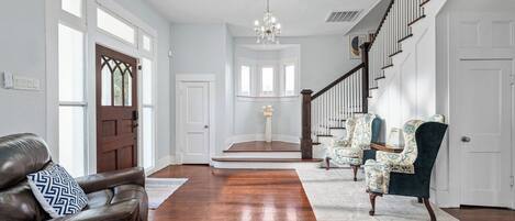 A spacious foyer with a wooden staircase, leather sofa, patterned armchairs, and a chandelier.