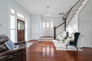A spacious foyer with a wooden staircase, leather sofa, patterned armchairs, and a chandelier. - NEW! Fall in a Victorian | Grand Porches Near DT (San Antonio)