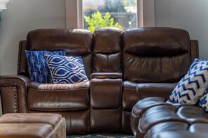 A brown leather sofa with a cushion, a side table, and a window with a view of greenery. - NEW! Fall in a Victorian | Grand Porches Near DT (San Antonio)