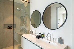Un baño moderno con cabina de ducha de vidrio, un tocador blanco con un lavamanos grande, dos espejos redondos y artículos de tocador sobre el lavamanos.