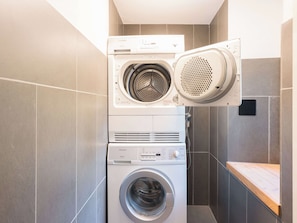 A stacked washer and dryer unit in a bathroom with tiled walls and a wooden bench.