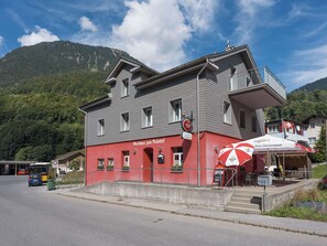 A building with a red facade and a balcony, located near a road with a bus and a trash bin.