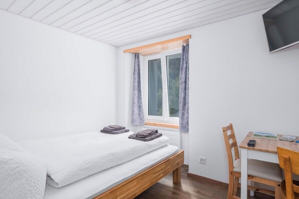 A bedroom with a bed, a window with curtains, a desk with a chair, and a television mounted on the wall.