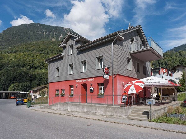 Un bâtiment à la façade rouge, doté d’un balcon, situé près d’une route où passe un autobus et où se trouve une poubelle.