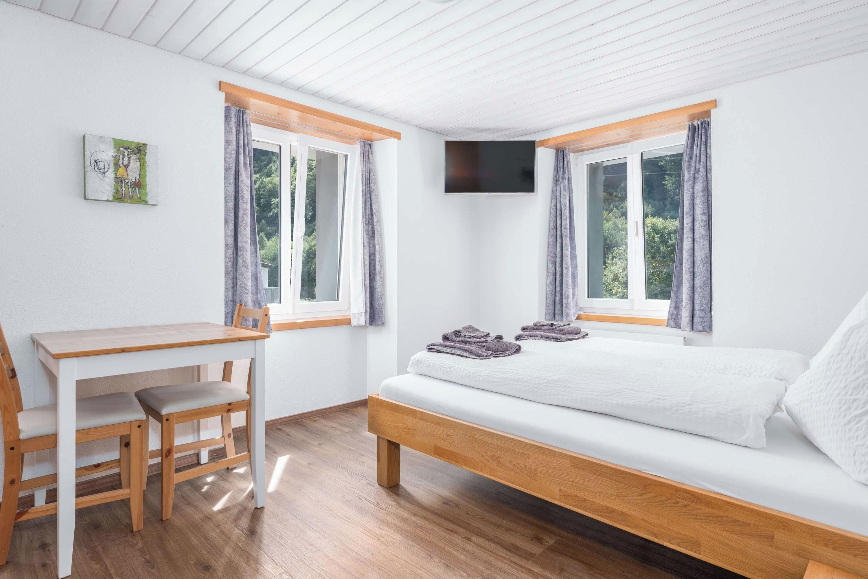 Une chambre à coucher avec un lit, un bureau et une chaise. Il y a deux fenêtres avec des rideaux.