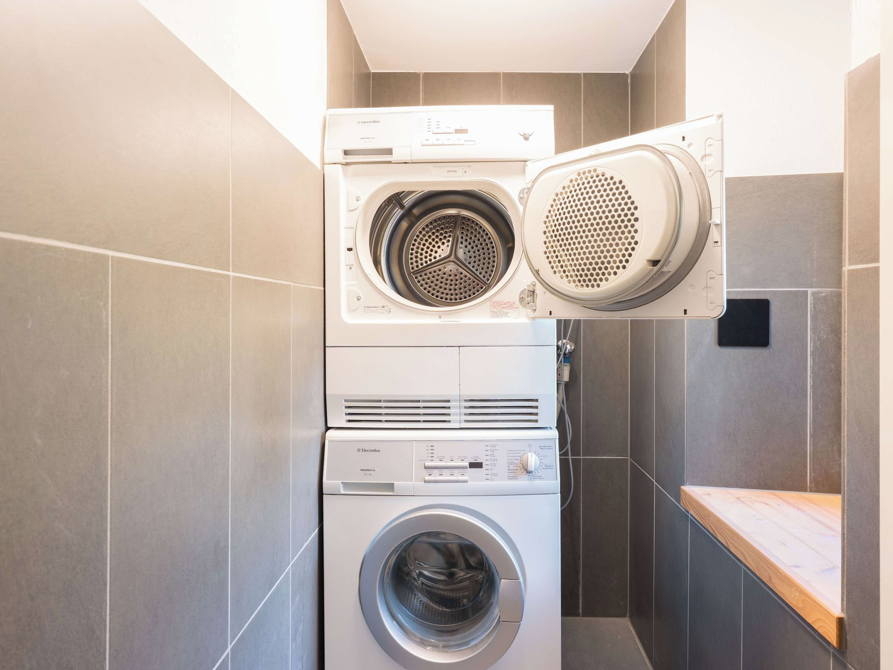 Une machine à laver et un sèche-linge superposés dans une salle de bain aux murs carrelés, avec un banc en bois.