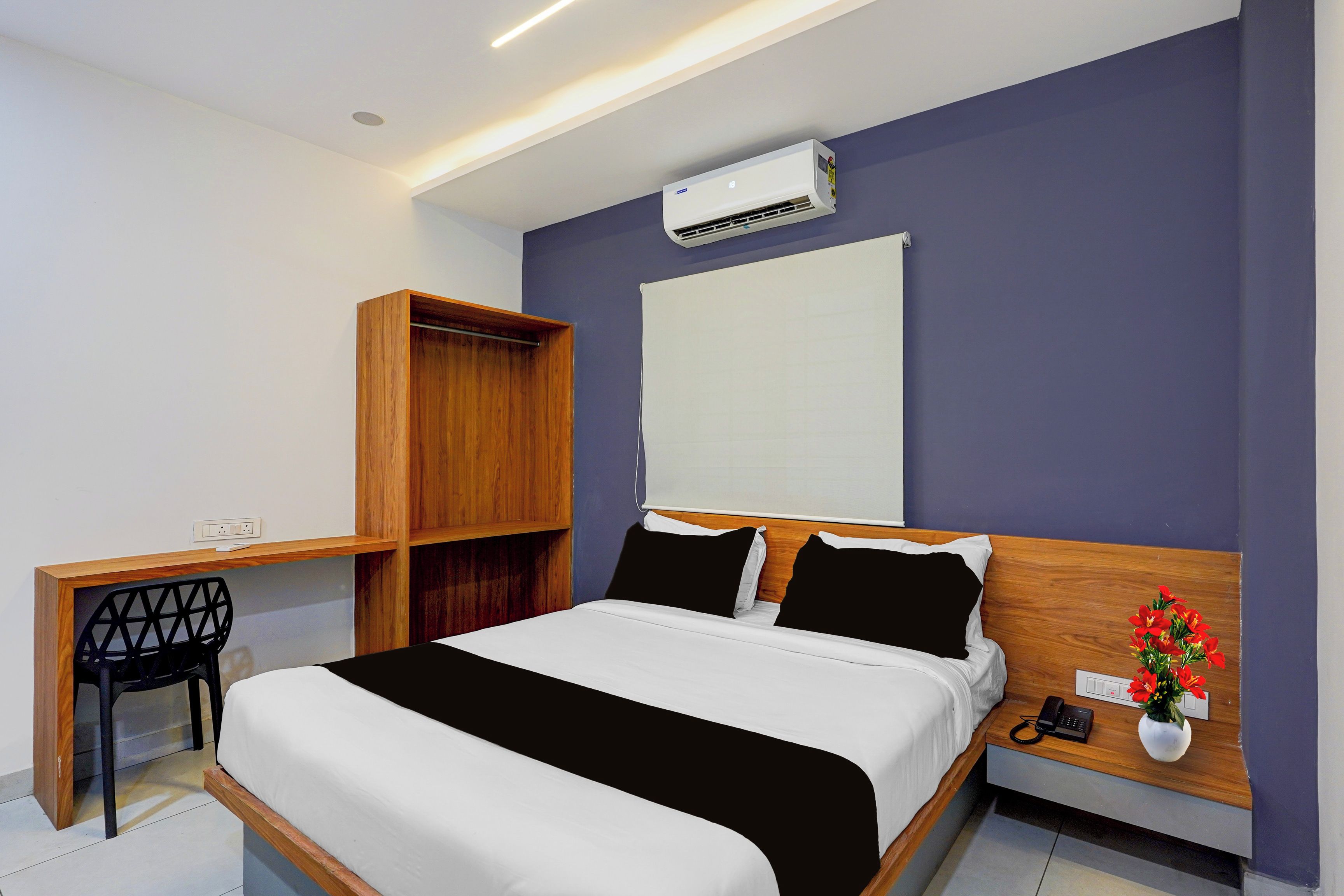A hotel room with a bed, a desk, a wardrobe, an air conditioning unit, a telephone, and a vase with flowers.