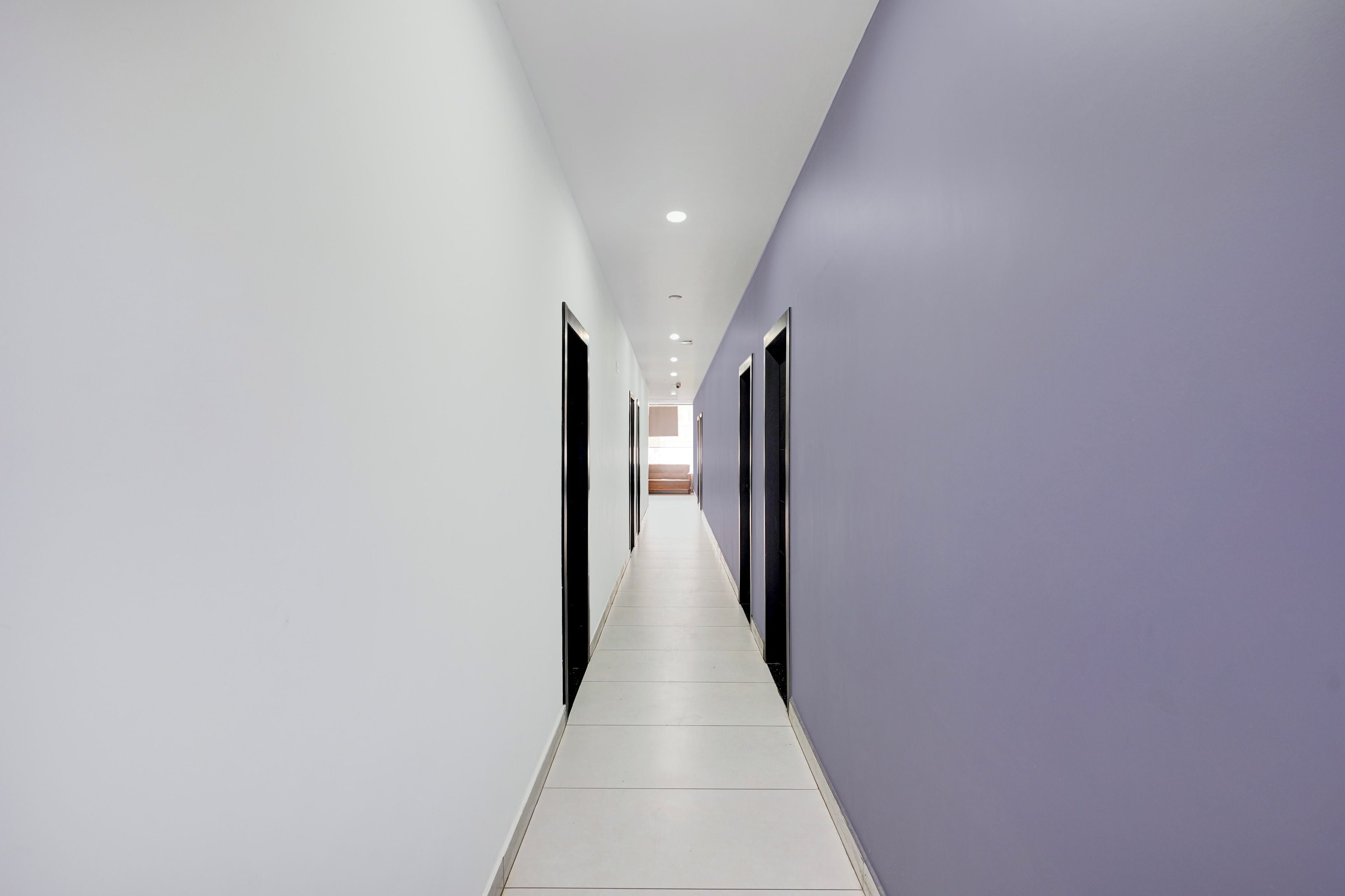 A long, narrow hallway with white and purple walls, leading to a room with a large window.
