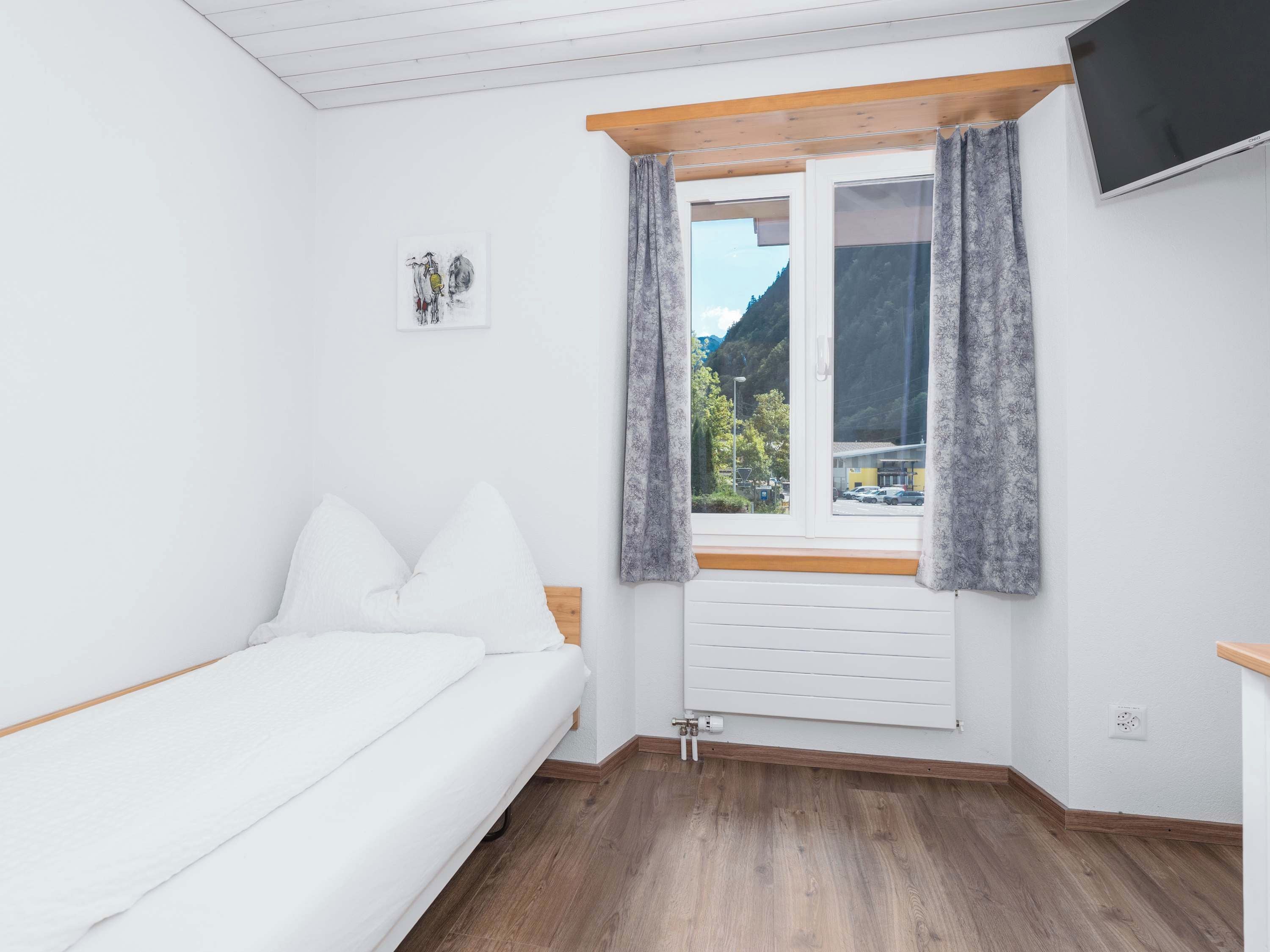 A bedroom with a bed, a window with curtains, a wall-mounted TV, and a radiator.