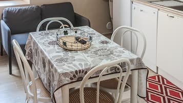 Zona pranzo con tavolo imbandito per quattro persone, un divano e angolo cottura con frigorifero e pensili.