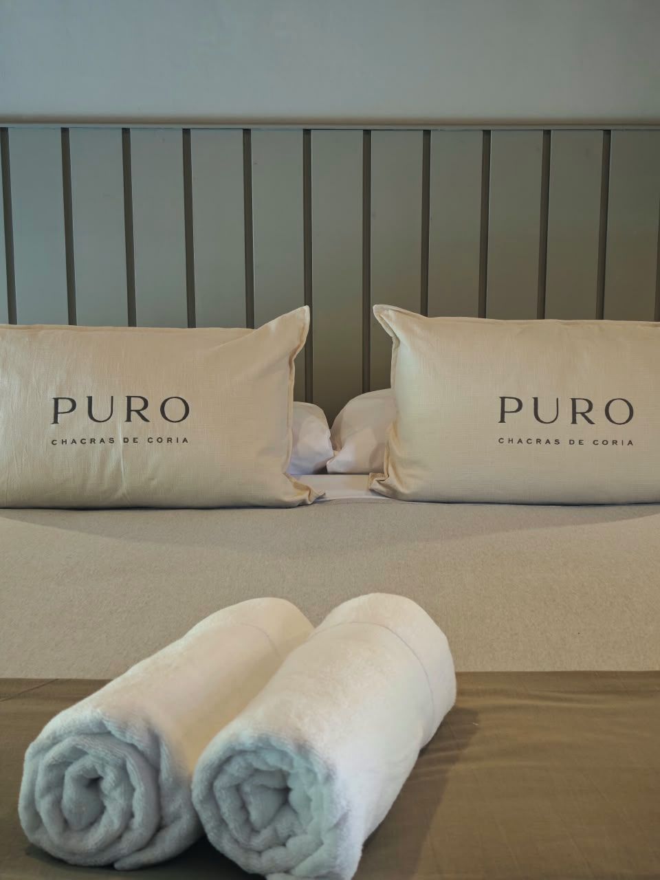 Ein Bett mit zwei Kissen, beschriftet mit 'PURO CHACRAS DE CORIA', und zwei gerollten Handtüchern auf dem Bett.
