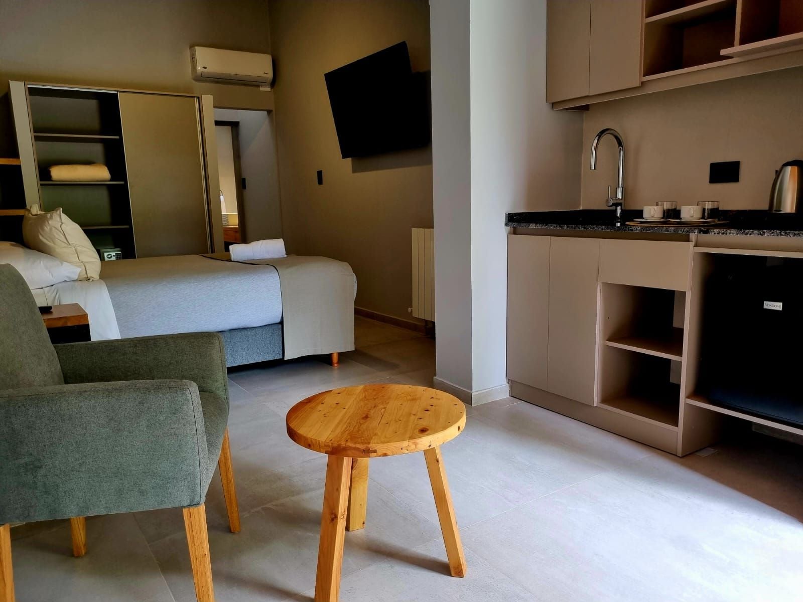A compact hotel room with a bed, a seating area, a small wooden table, a kitchenette, and a flat-screen TV mounted on the wall.
