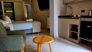 A compact hotel room with a bed, a seating area, a small wooden table, a kitchenette, and a flat-screen TV mounted on the wall.