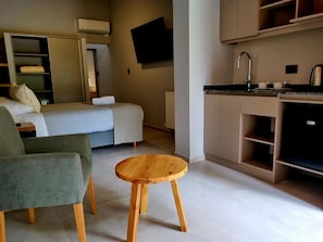 Habitación de hotel compacta con cama, zona de estar, una pequeña mesa de madera, kitchenette y televisor de pantalla plana montado en la pared.