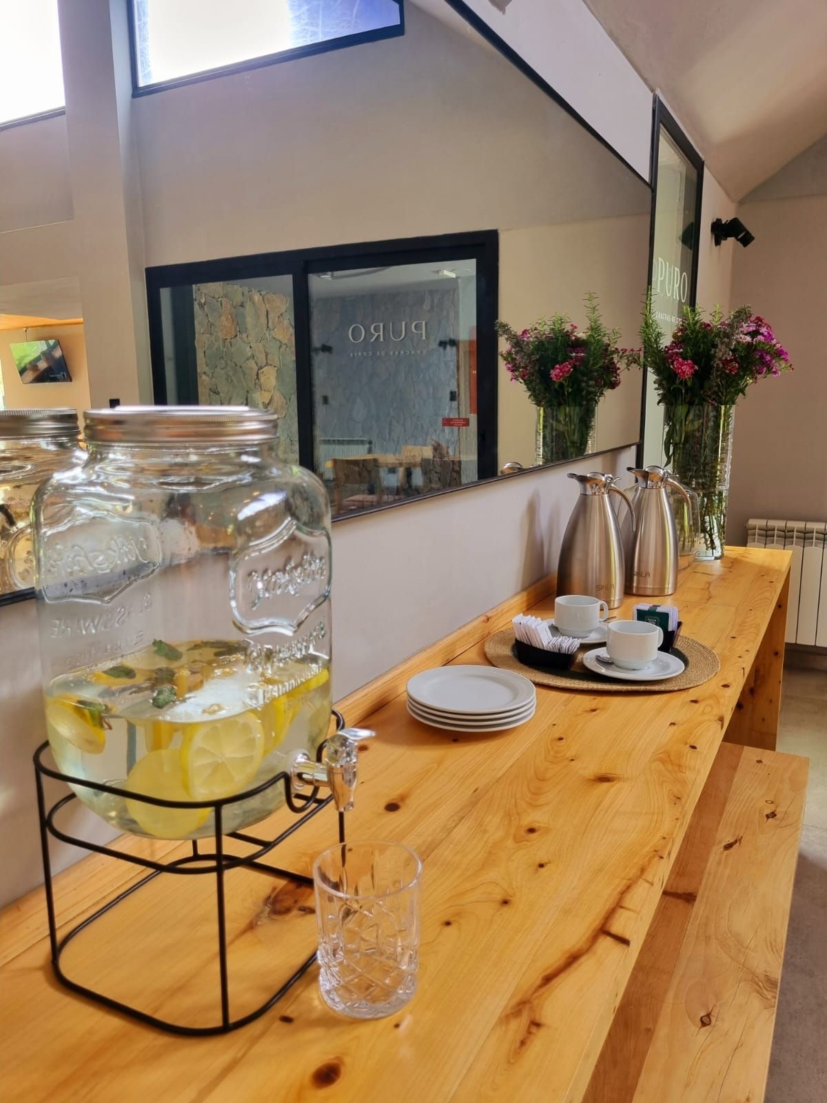 Una mesa de madera con un tarro de vidrio grande con agua, un vaso y una jarra metálica. Hay platos, tazas y una cesta pequeña sobre la mesa.