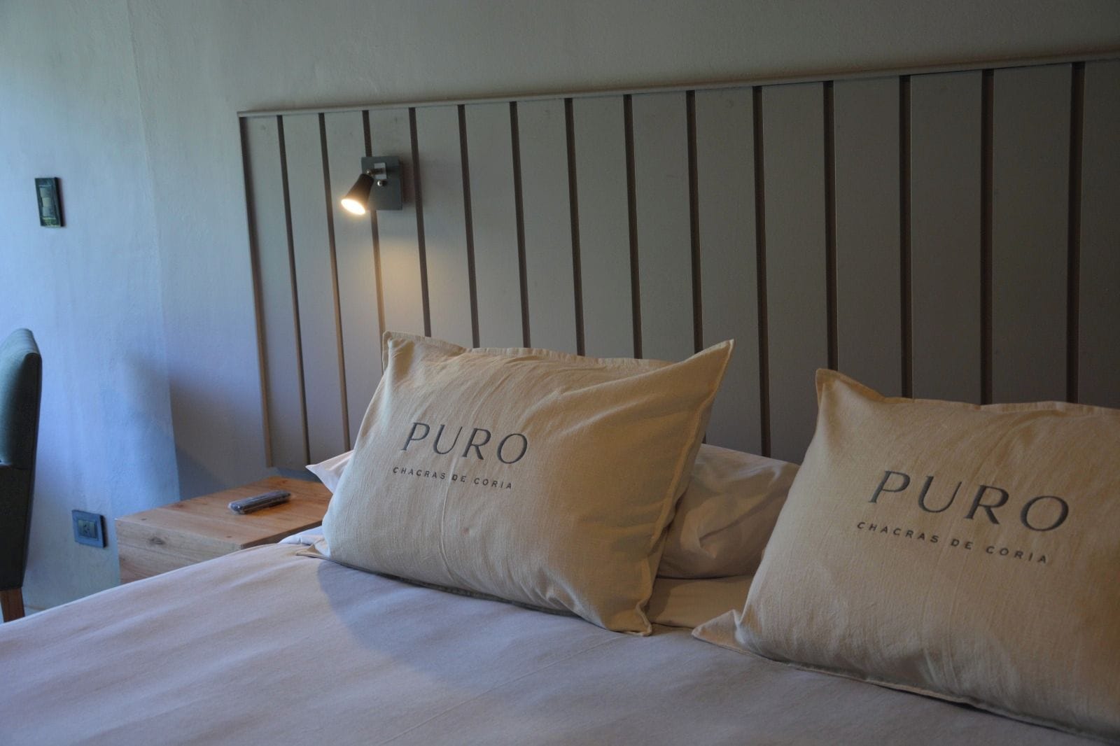 Una habitación de hotel con una cama, dos almohadas con el texto 'PURO CHACRAS DE CORIA', una mesita de noche, un control remoto y una lámpara de pared.