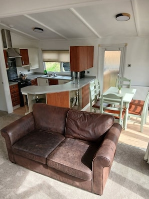 A living room with a brown leather sofa, a dining area with a table and chairs, and a kitchen with wooden cabinets and a stove.