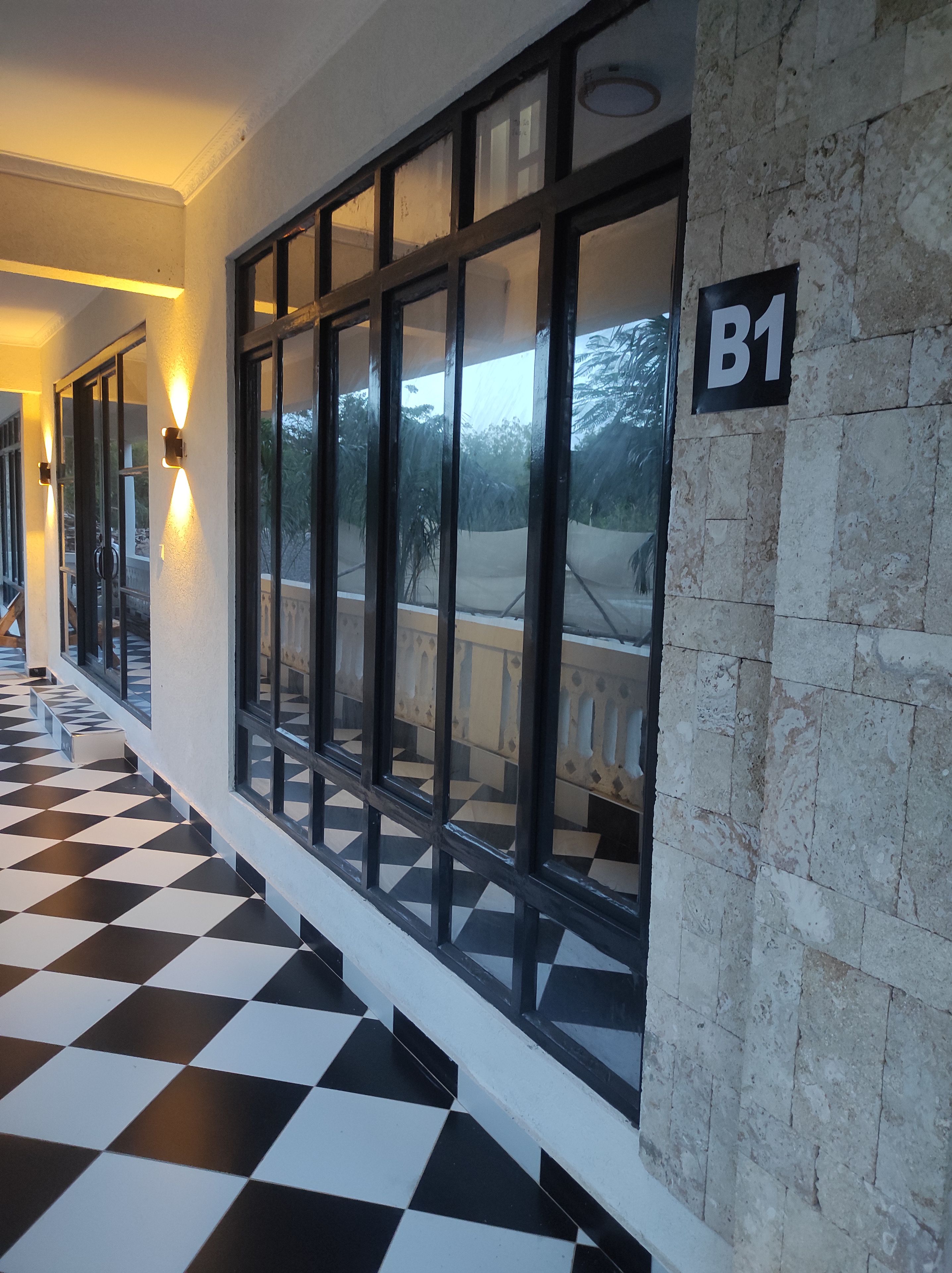 A corridor with a checkered floor, a wall-mounted sign reading 'B1', and a window with a view of greenery.