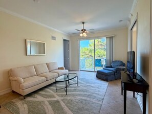 Una sala de estar con un sofá color beige, una mesa de centro de vidrio, un televisor sobre un mueble bajo y un balcón con vistas a árboles.