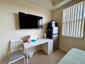 A compact room with a white desk, a chair, a bed, a TV mounted on the wall, and a window with blinds. - Bright Studio - Gulf Access - Bonita Springs (Bonita Springs)