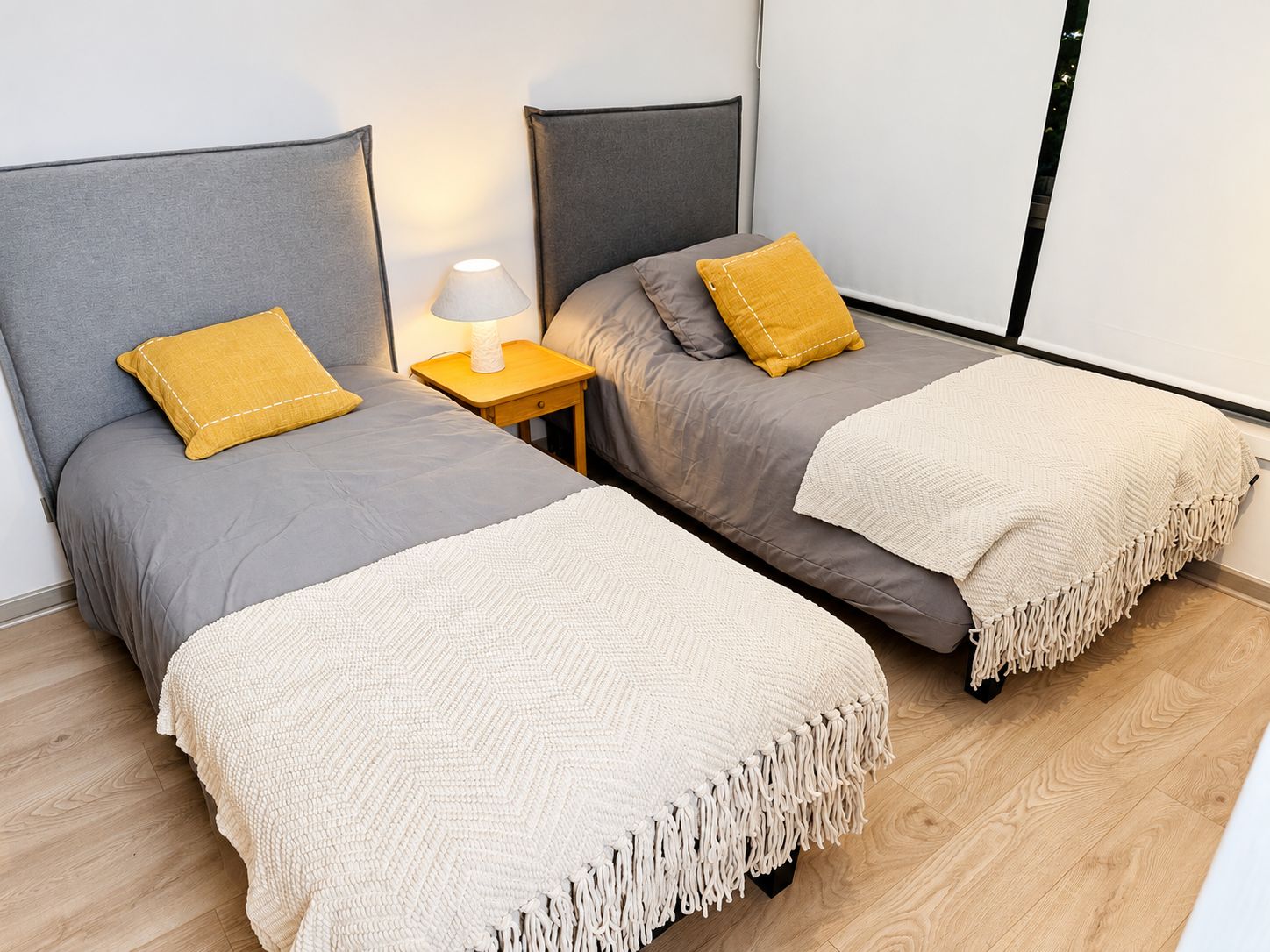 A double bed with a grey headboard and a yellow pillow, a bedside table with a lamp, and a beige throw blanket.
