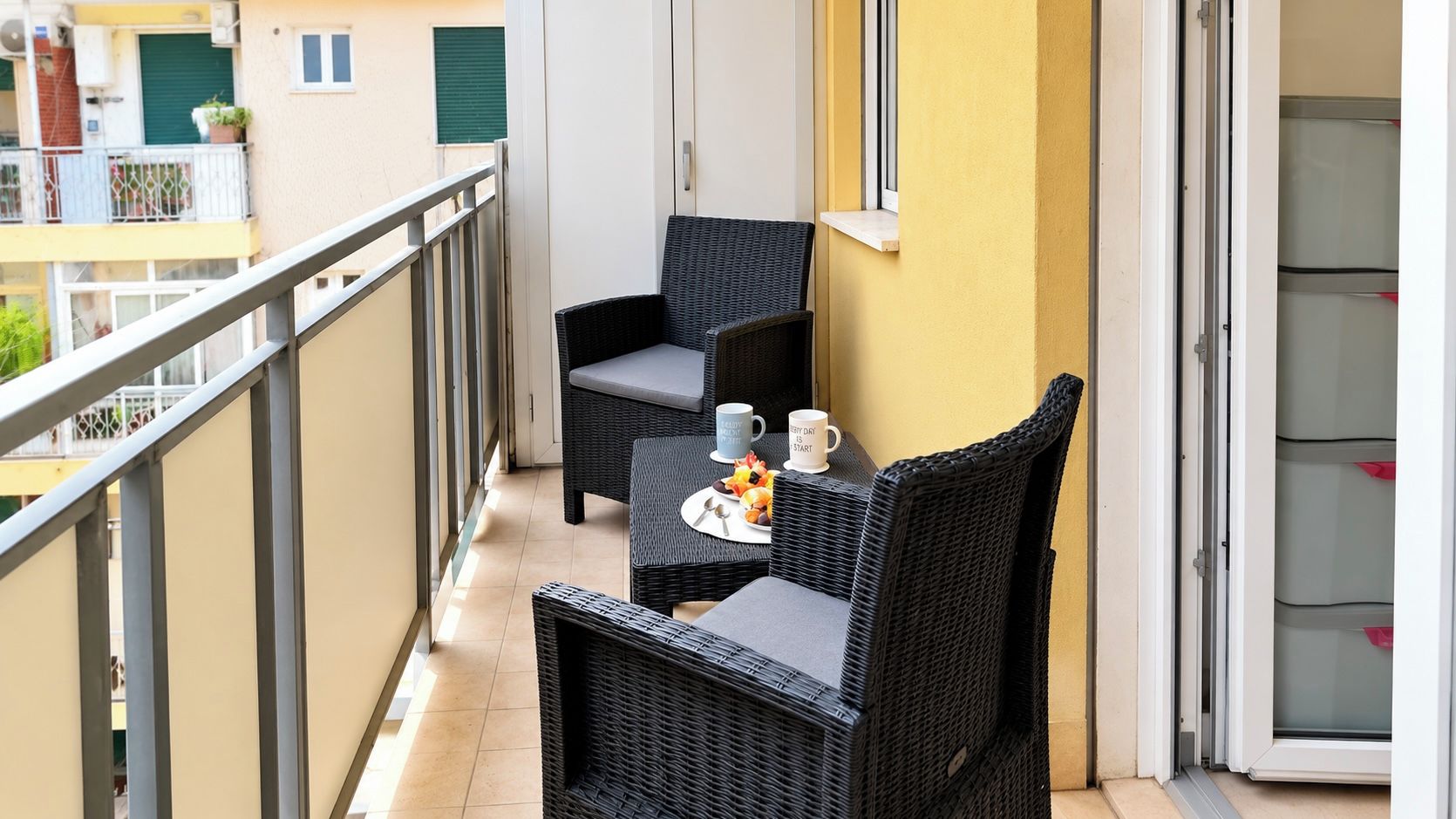 A balcony with wicker furniture, a small table with a cup and fruits, and a view of a building with balconies.