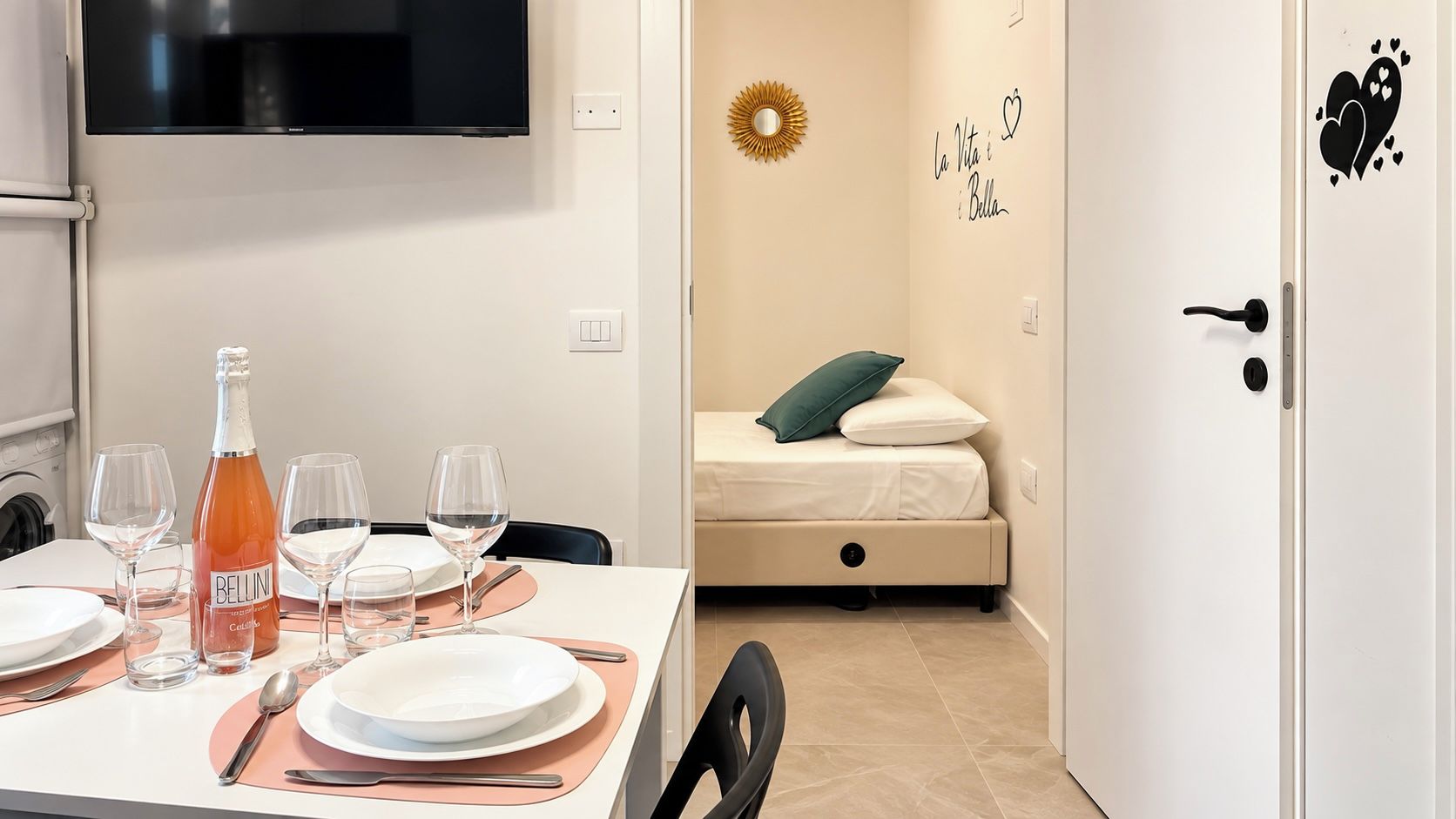 A neatly arranged dining area with a table set for four, a bottle of Bellini wine, and four glasses. A bed with a green pillow is visible in the background.