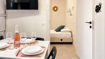 A neatly arranged dining area with a table set for four, a bottle of Bellini wine, and four glasses. A bed with a green pillow is visible in the background.