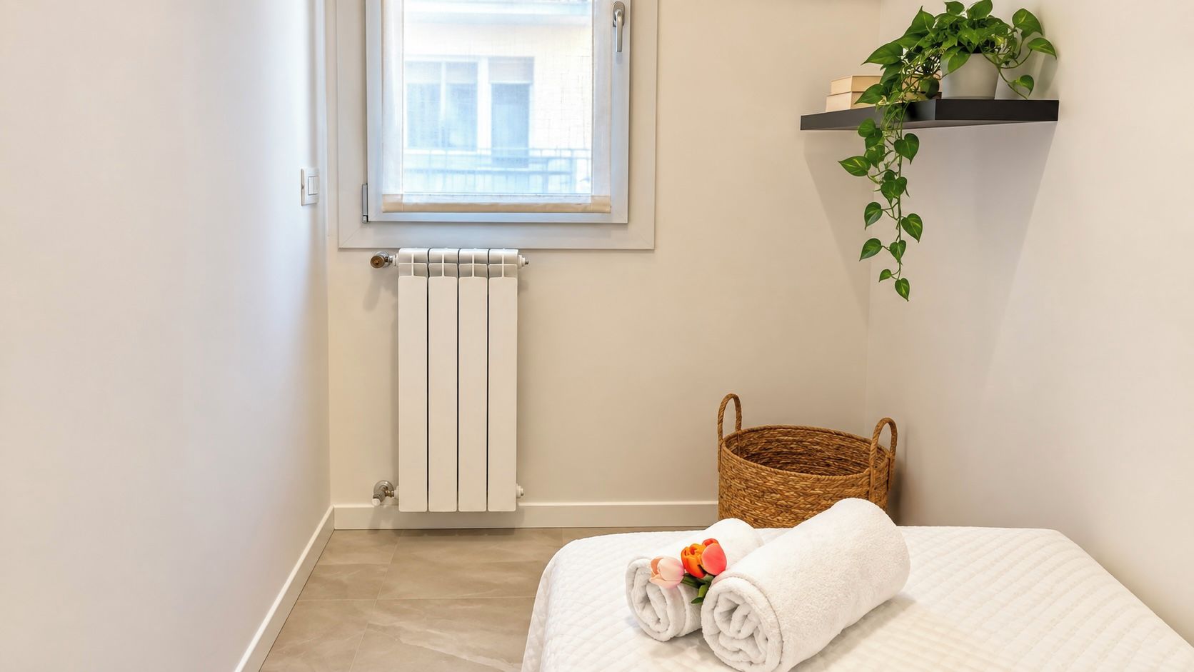 A hotel room with a bed, a radiator, a window, a plant, and a basket.