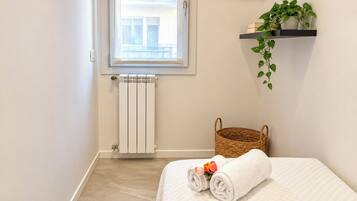 A hotel room with a bed, a radiator, a window, a plant, and a basket.