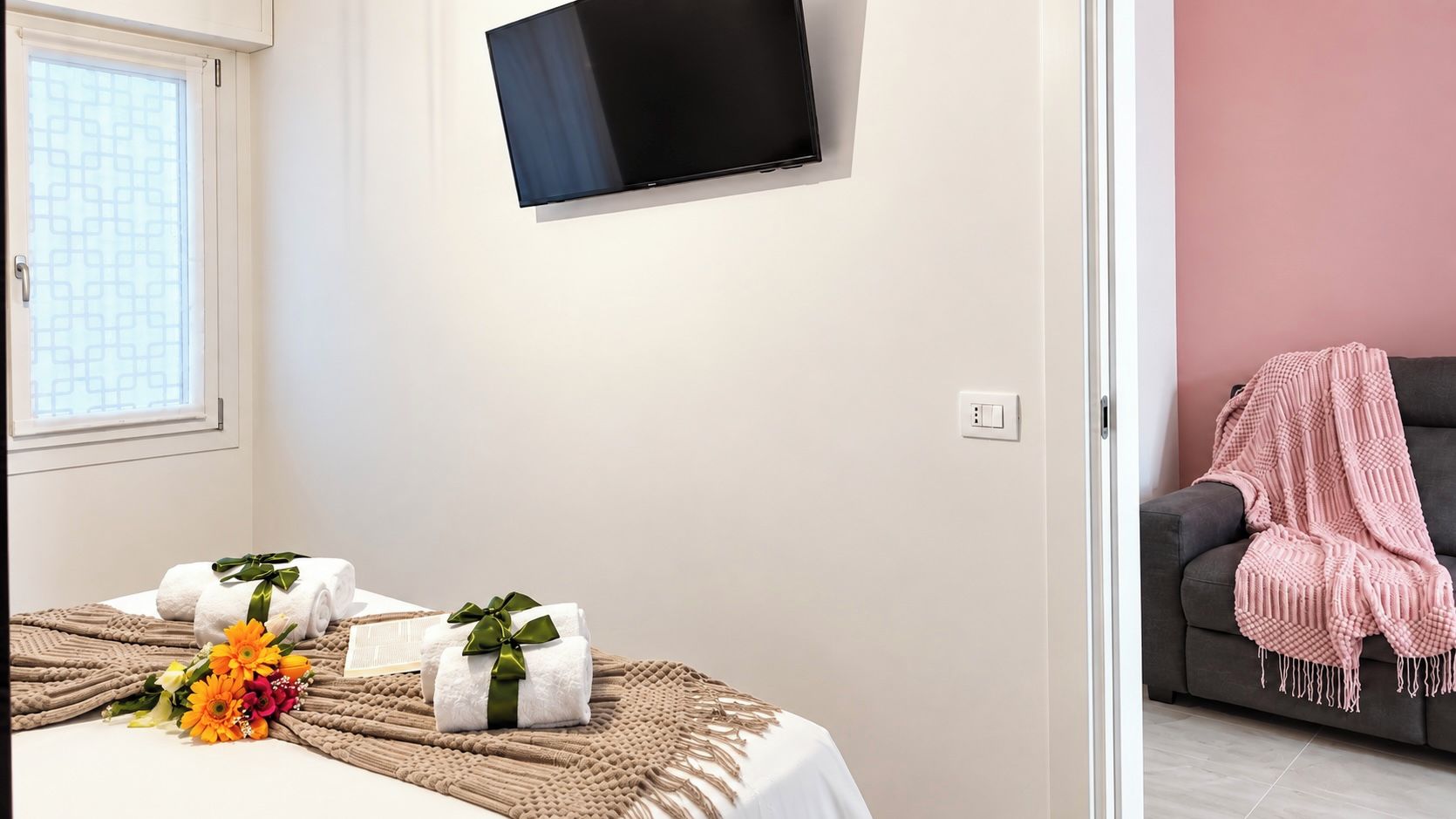 A hotel room with a bed, a TV mounted on the wall, a window with a view, and a sofa with a blanket.
