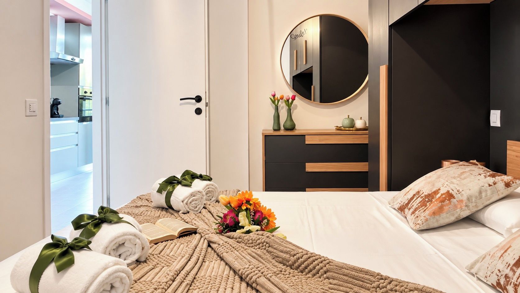 A hotel room with a bed, a bedside table with a mirror, a small dresser, and a view of the kitchen through an open door.