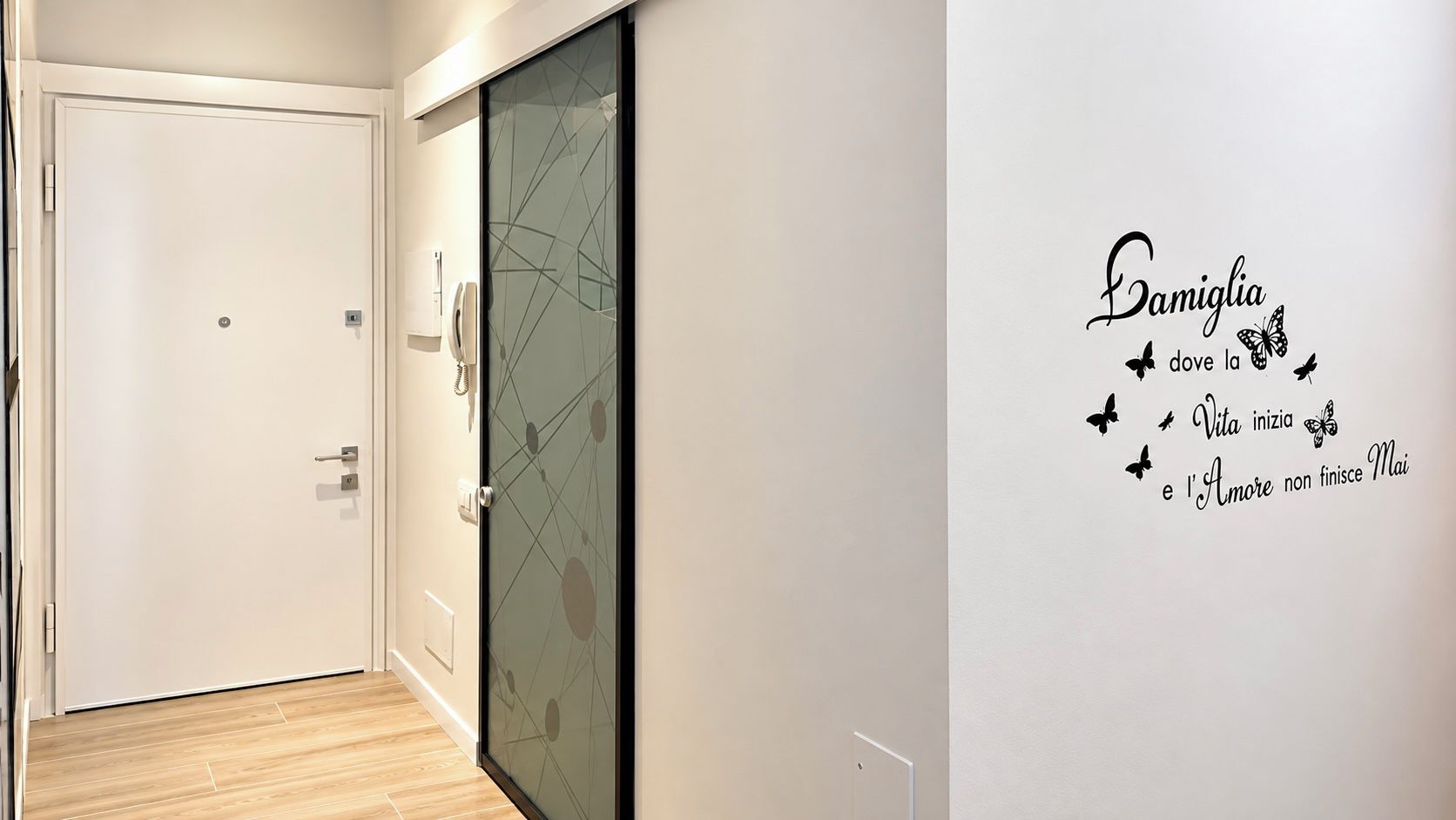A hallway with a glass door, a white wall with a decorative phrase, and a wooden floor.