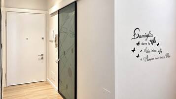 A hallway with a glass door, a white wall with a decorative phrase, and a wooden floor.