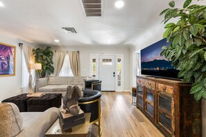 Ein Wohnzimmer mit einer Couch, Sesseln, einem Couchtisch, einem Fernseher auf einer Holzkommode, einer Pflanze und einer Tür mit Fenster.