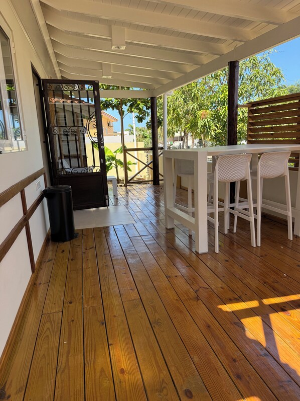 A covered outdoor area with a wooden floor, a white table with two chairs, and a view of the street and greenery.