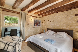 A bedroom with a stone wall, a bed with white bedding, a wooden headboard, a bedside table, a chair, and a window with curtains. - Room 'Chambre Lune' with Shared Pool, Wi-Fi and Air Conditioning - Table d'Hôtes (Salignac Eyvigues)