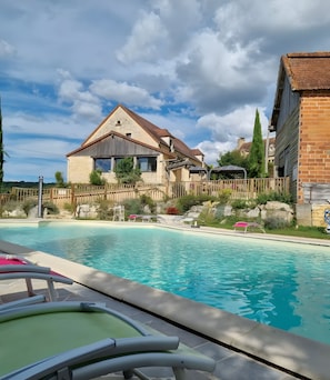 Ein Schwimmbecken mit Liegestühlen, einem Holzzaun und einem Steinhaus im Hintergrund.