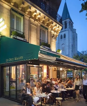Ein Straßencafé mit Tischen und Stühlen, ein Gebäude mit einer grünen Markise und ein Kirchturm im Hintergrund.