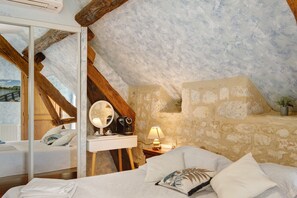 Une chambre avec un mur en pierre, des poutres apparentes, un lit avec une literie blanche, une table de chevet avec une lampe et une fenêtre donnant sur un plan d’eau.